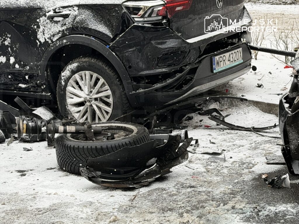 Zniszczony czarny samochód osobowy leży w śniegu, obok znajduje się oderwane koło i resztki zawieszenia.