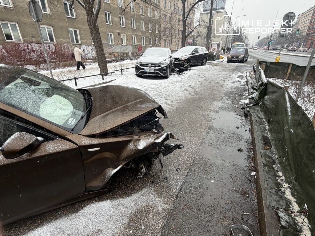 Zniszczony samochód osobowy na zaśnieżonej ulicy, obok stoją inne pojazdy, w tle przechodzień.