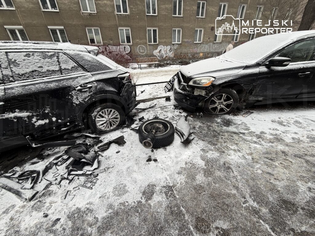 Dwa uszkodzone samochody leżą na zaśnieżonej ulicy, obok leżą fragmenty zderzaków i kół.