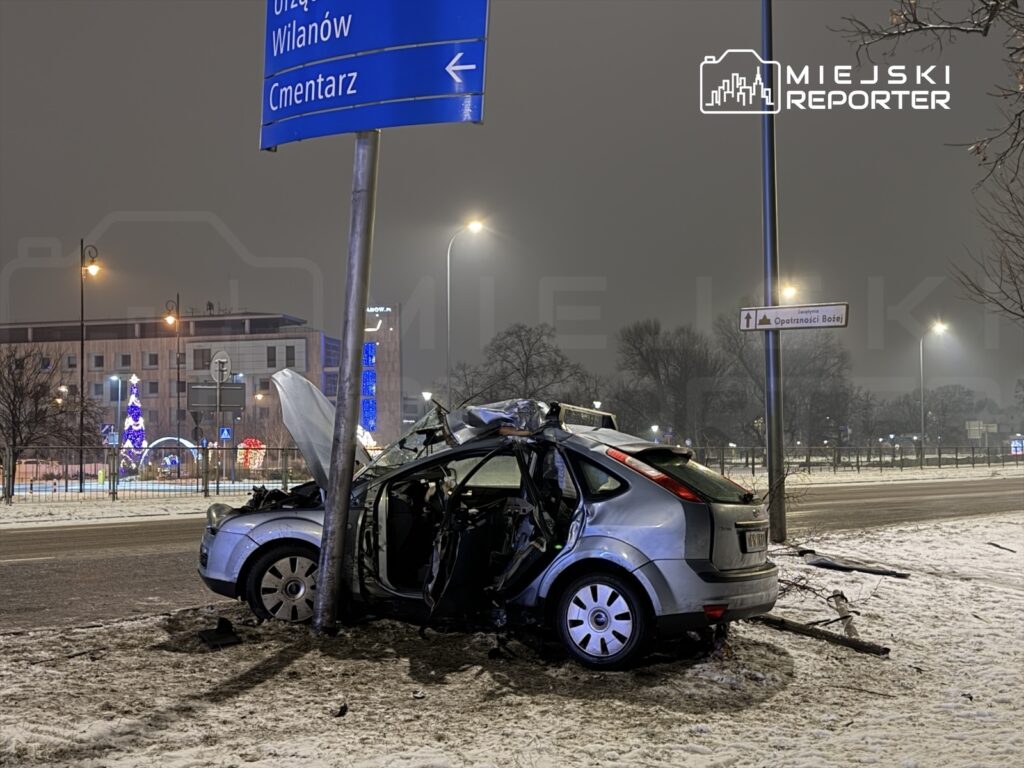Samochód osobowy uderzył w latarnię, z otwartą maską, na zaśnieżonej ulicy w nocy, obok świątecznych dekoracji.