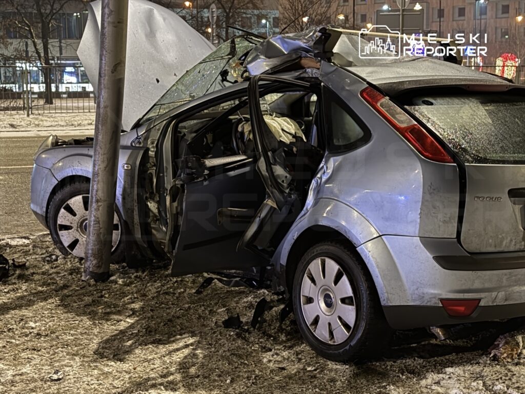 Rozbity samochód osobowy z otwartą maską stoi przy słupie, z widocznymi uszkodzeniami nadwozia.