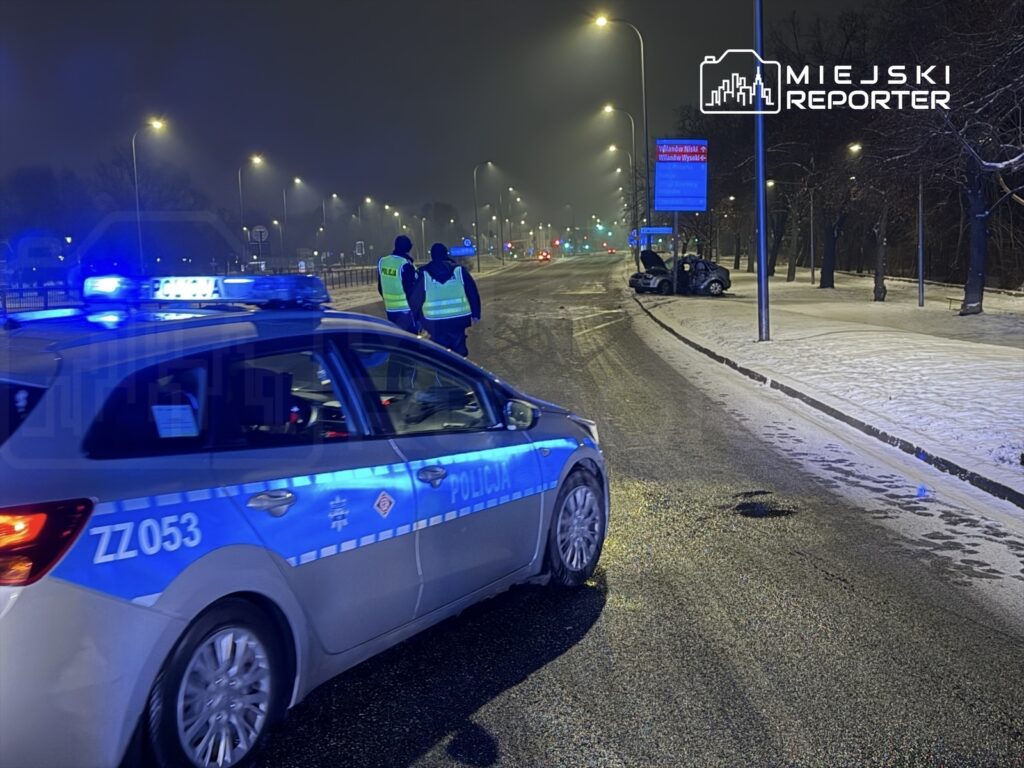 Policyjny radiowóz z włączonymi sygnałami świetlnymi zatrzymany na ośnieżonej drodze, obok dwóch funkcjonariuszy rozmawiających.