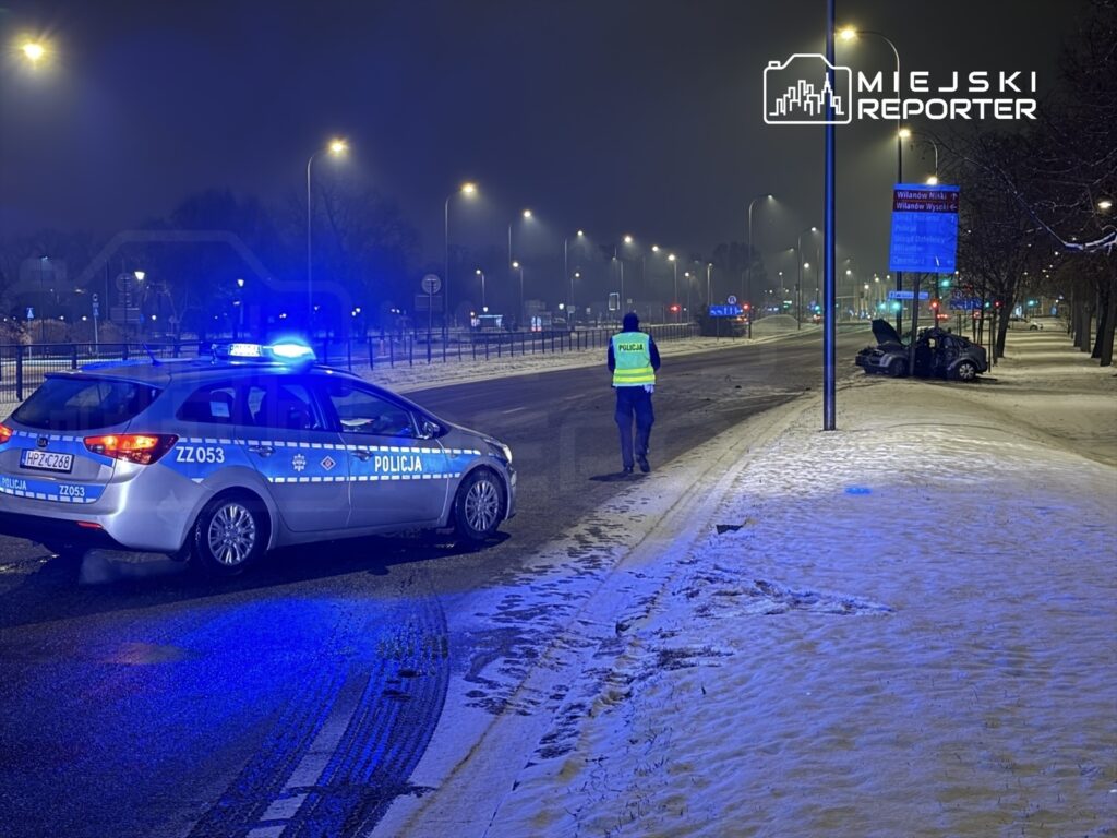 Policyjny radiowóz z włączonymi sygnałami świetlnymi stoi na zaśnieżonej drodze, a funkcjonariusz w kamizelce idzie w stronę uszkodzonego samochodu.