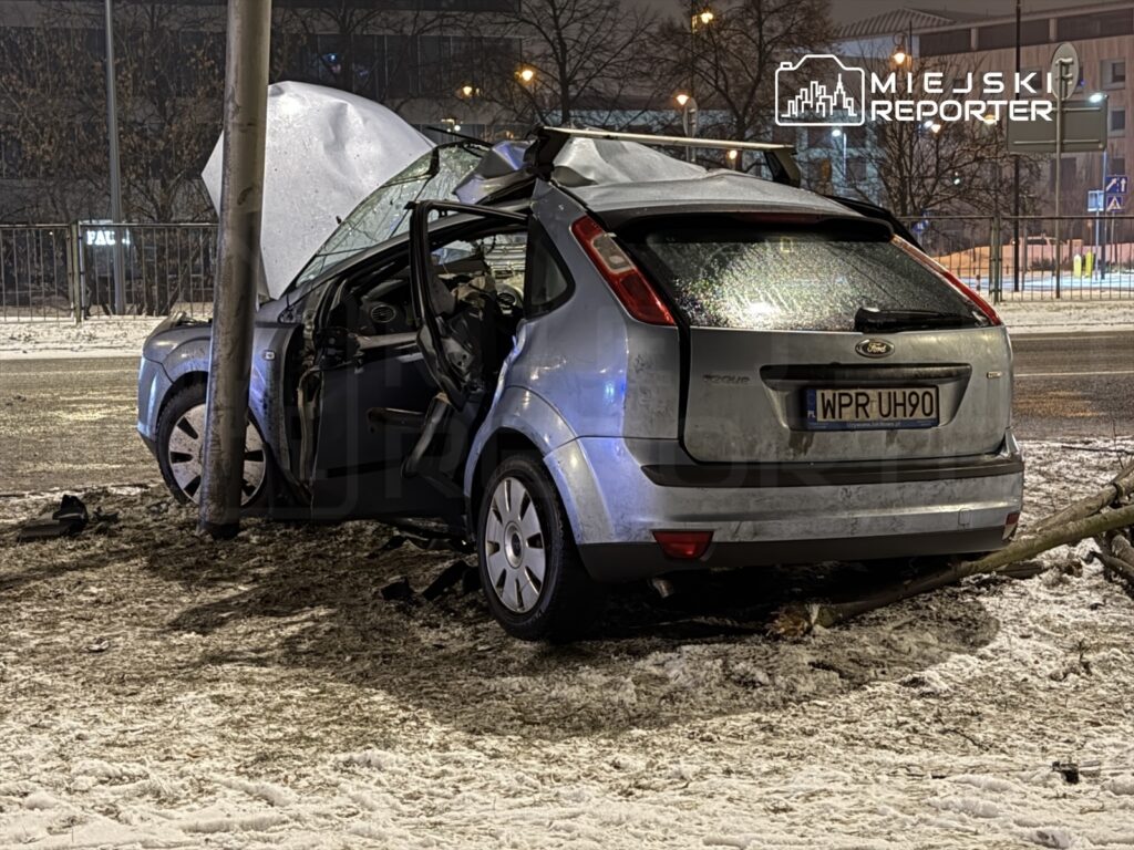 Uszkodzony samochód osobowy stoi przy latarni, z otwartymi drzwiami na zaśnieżonej ulicy nocą.