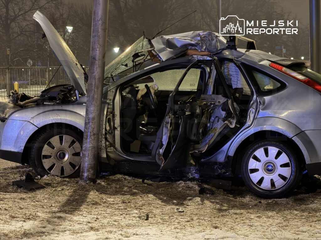 Zniszczony samochód osobowy z otwartą maską stoi przy latarni, obok widać uszkodzone drzwi i śnieg na ziemi.