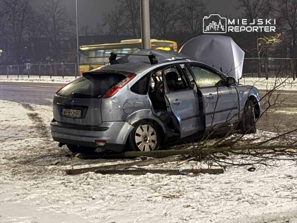 Zniszczony samochód osobowy leży na poboczu drogi, obok niego leży połamana gałąź, a wokół jest pokryta śniegiem nawierzchnia.