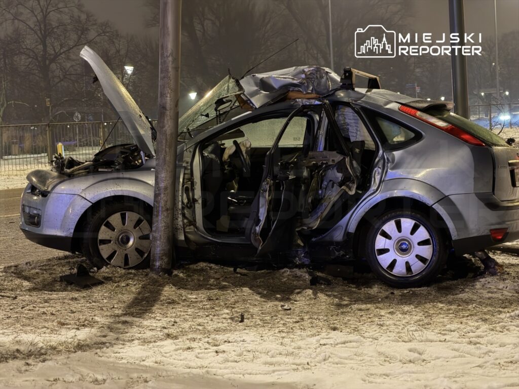 Zniszczony samochód osobowy uderzony w latarnię, z otwartą maską i uszkodzoną karoserią, na śnieżnej nawierzchni.