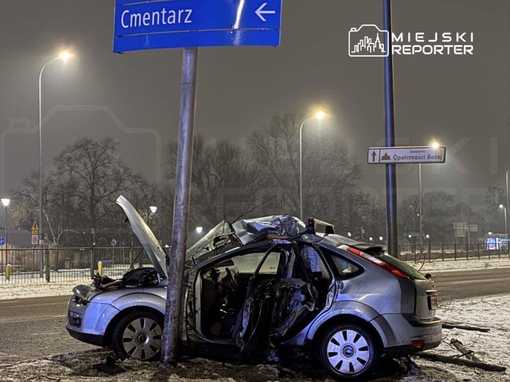 Zniszczony samochód osobowy stoi przy latarni, z otwartą maską, obok znaku kierunkowym do cmentarza.