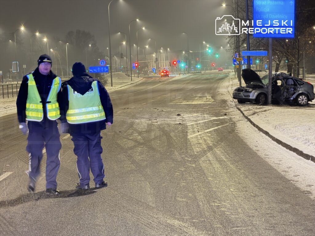 Dwaj policjanci w odblaskowych kamizelkach idą wzdłuż oświetlonej ulicy obok uszkodzonego samochodu.