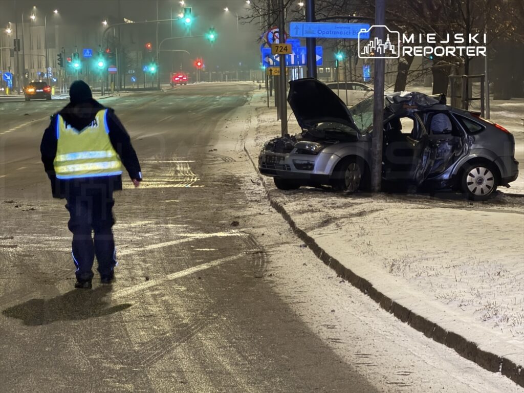 Policjant w odblaskowej kamizelce zmierza w kierunku uszkodzonego samochodu zaparkowanego przy ulicy w nocy.