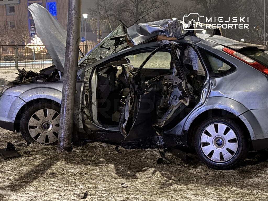 Zniszczony samochód osobowy uderzony w latarnię, z otwartą maską i uszkodzoną boczną częścią.