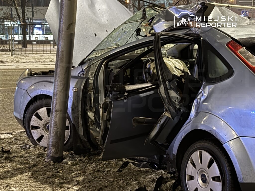 Samochód osobowy zniszczony w wyniku kolizji z latarnią, otwarte drzwi, widoczne uszkodzenia nadwozia.