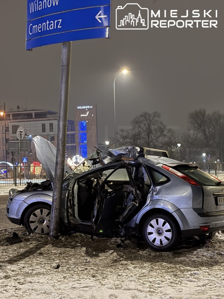 Zniszczony samochód osobowy stoi przy drodze, uderzony w znak drogowy, na tle oświetlonego miasta nocą.