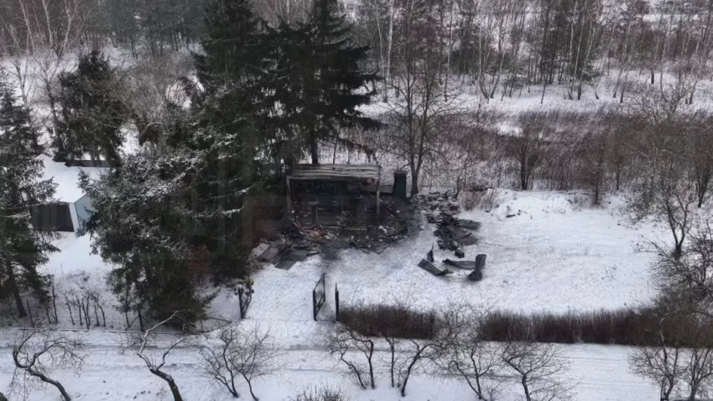 Zniszczony budynek otoczony drzewami, pokryty śniegiem, z pozostałościami materiałów budowlanych w tle.