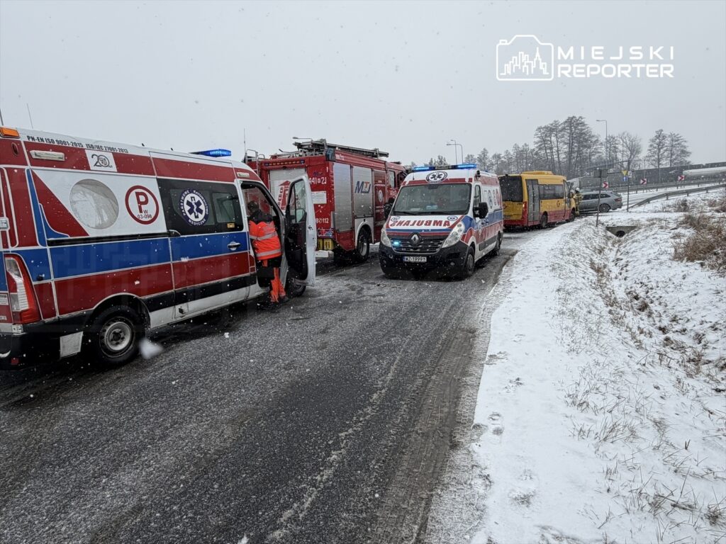 Zespół ratownictwa medycznego pracuje na zaśnieżonej drodze przy wypadku, obok pojazdów straży pożarnej i autobusu.
