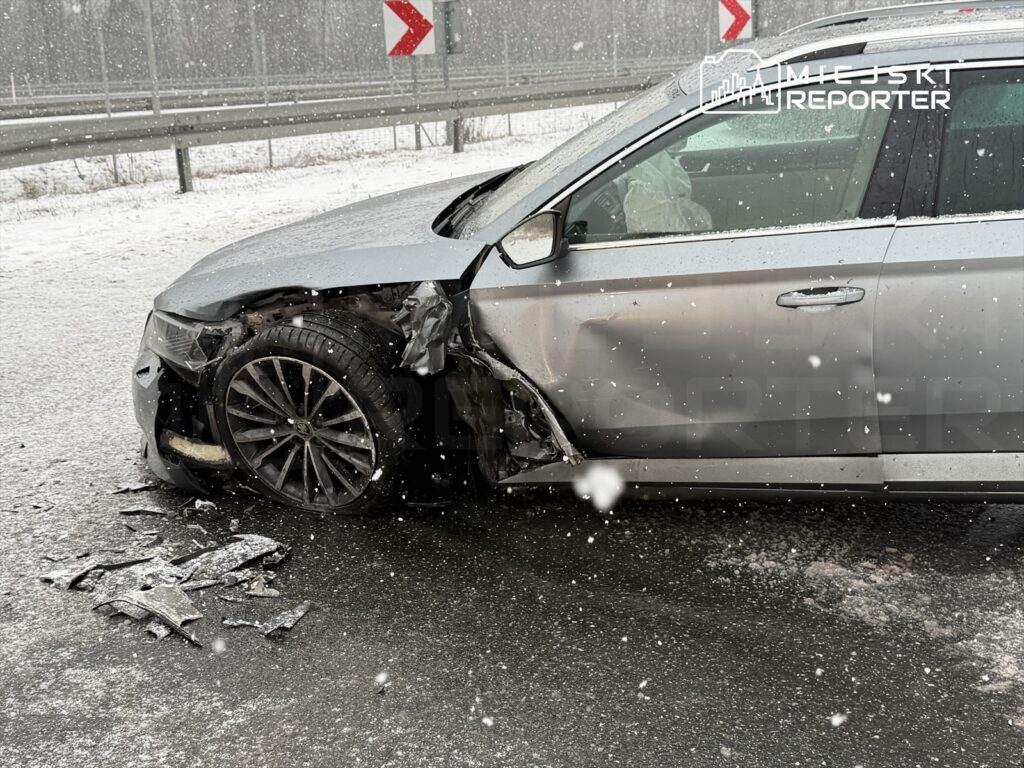 Uszkodzony samochód osobowy stoi na drodze w trakcie opadów śniegu, z widocznymi uszkodzeniami nadwozia.