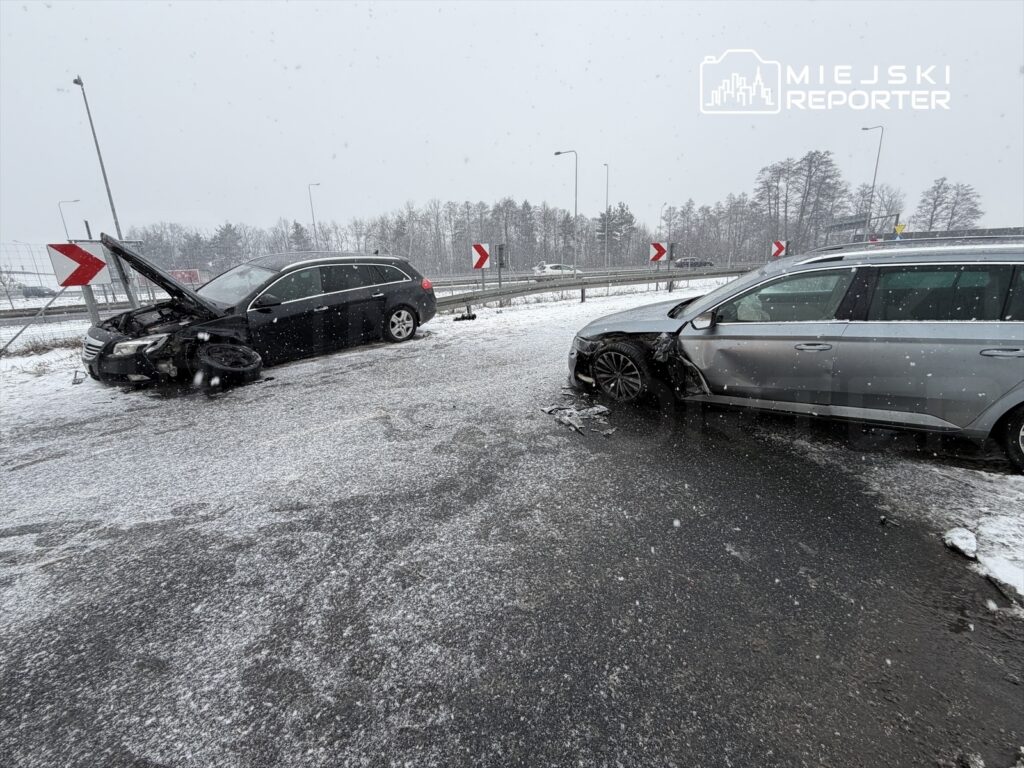 Dwa uszkodzone samochody na zaśnieżonej drodze, jeden z otwartą maską, w pobliżu znaku kierunkowego.