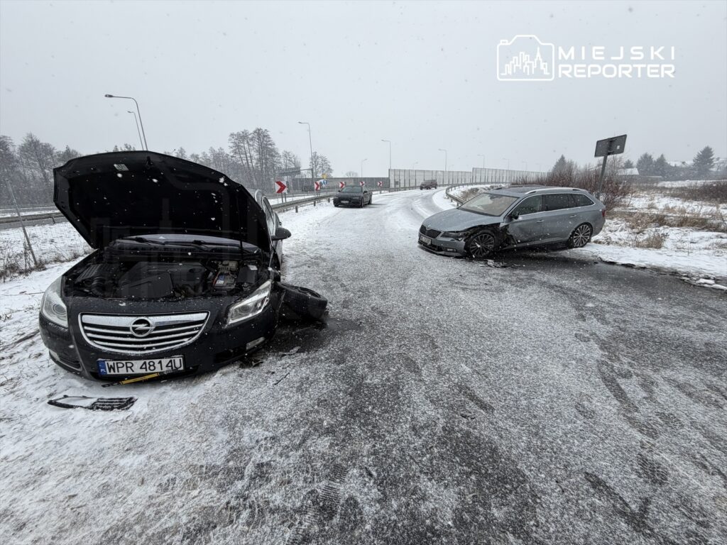 Dwa uszkodzone samochody na ośnieżonej drodze, jeden z otwartą maską, w tle widoczne znaki drogowe i zaspy śnieżne.
