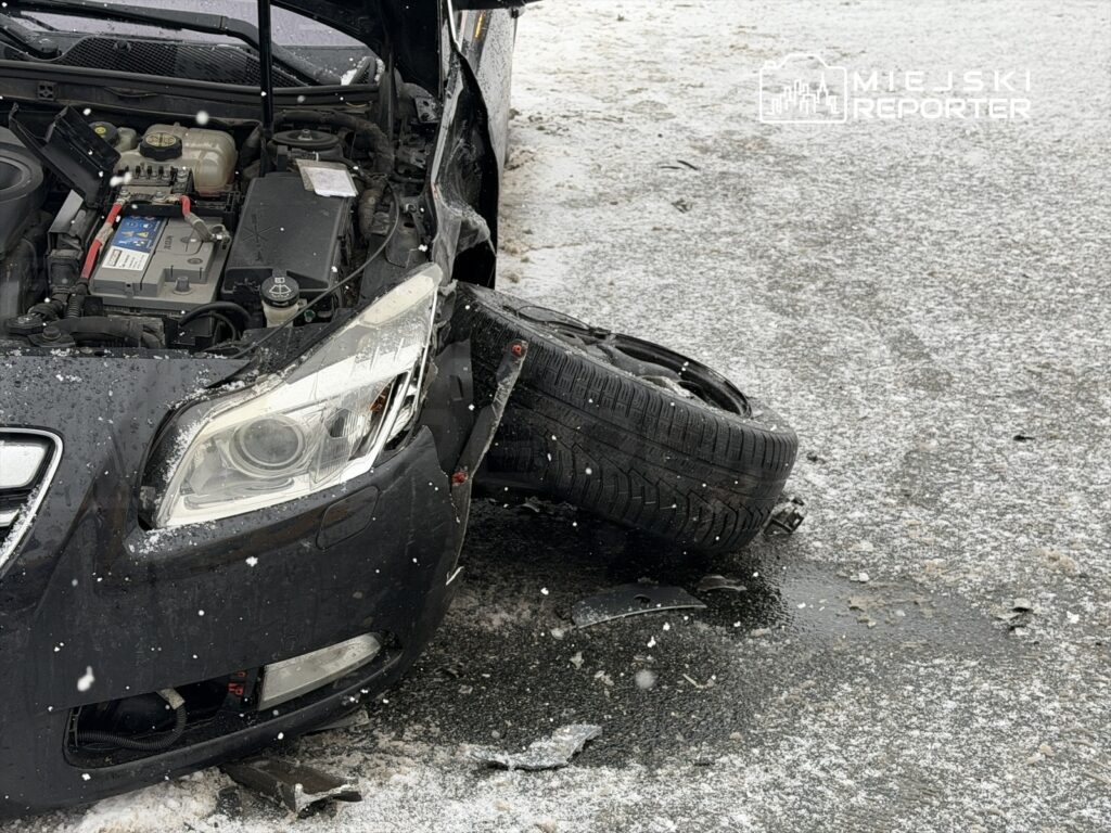 Czarny samochód osobowy z uszkodzoną przednią częścią i odczepionym kołem leży na śnieżnej nawierzchni.