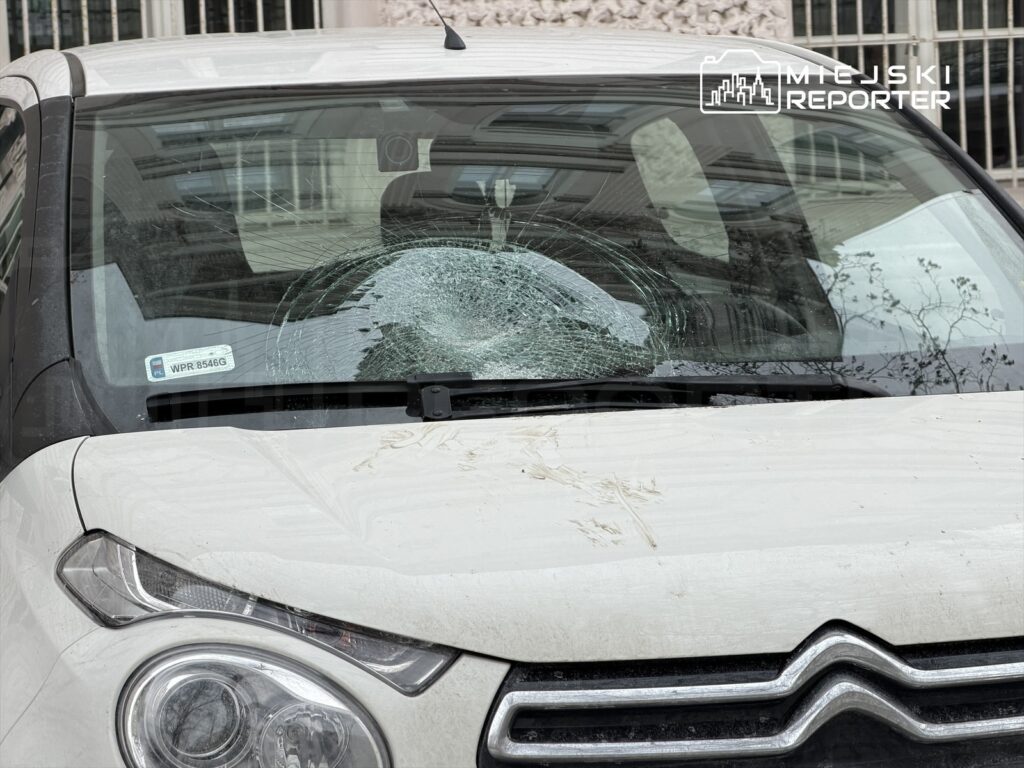 Uszkodzona szyba czołowa białego samochodu osobowego z widocznymi pęknięciami i śladami brudu.