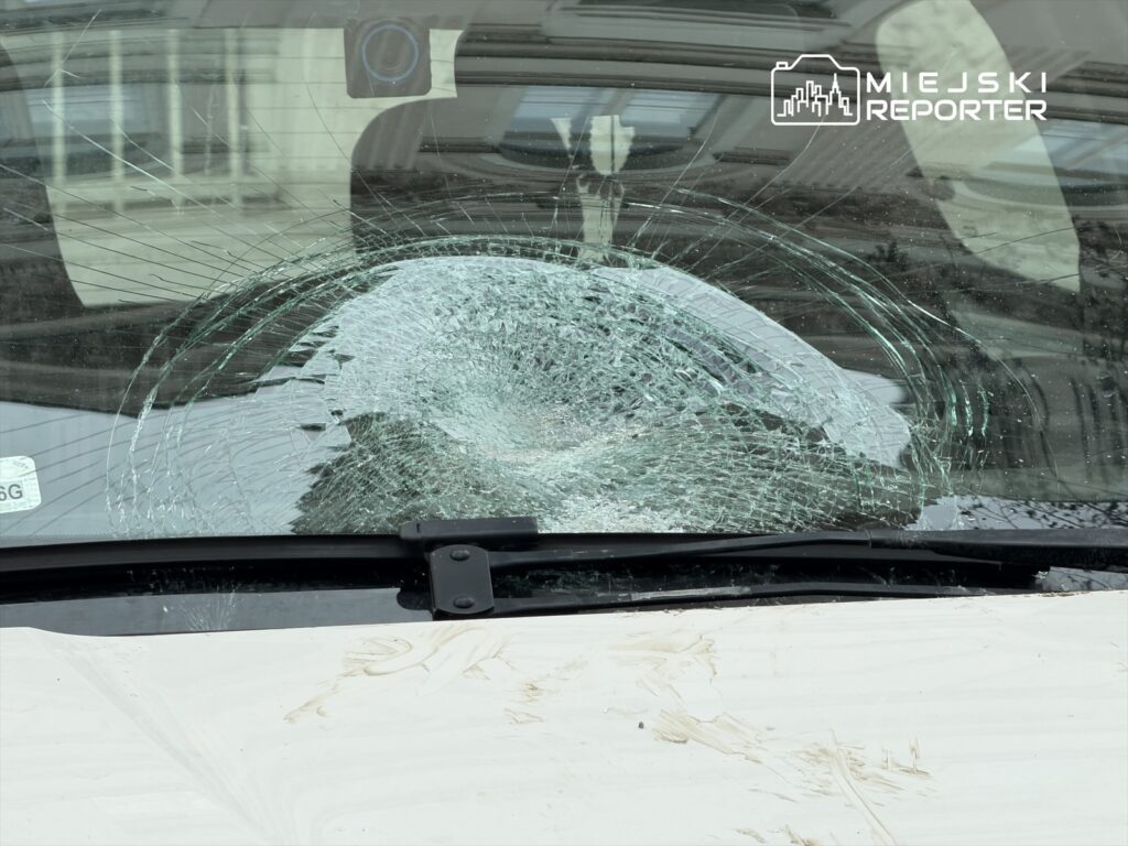 Zbita szyba przednia samochodu z wyraźnymi pęknięciami, znajdująca się na parkingu.
