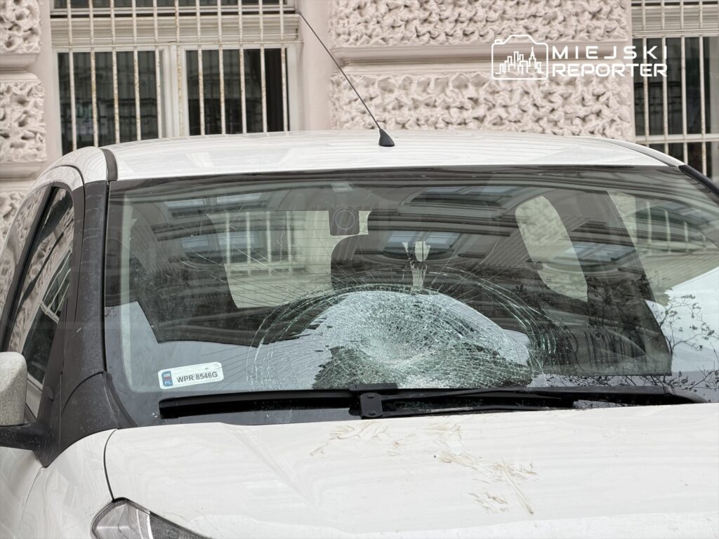 Pęknięta szyba czołowa białego samochodu zaparkowanego przy ozdobnym budynku.