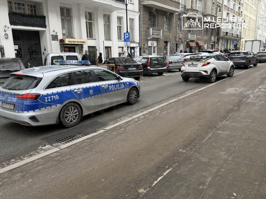 Policyjny radiowóz zaparkowany na ulicy, obok stojące samochody, w tle widoczny budynek z napisem "Pizza przy Politechnice".