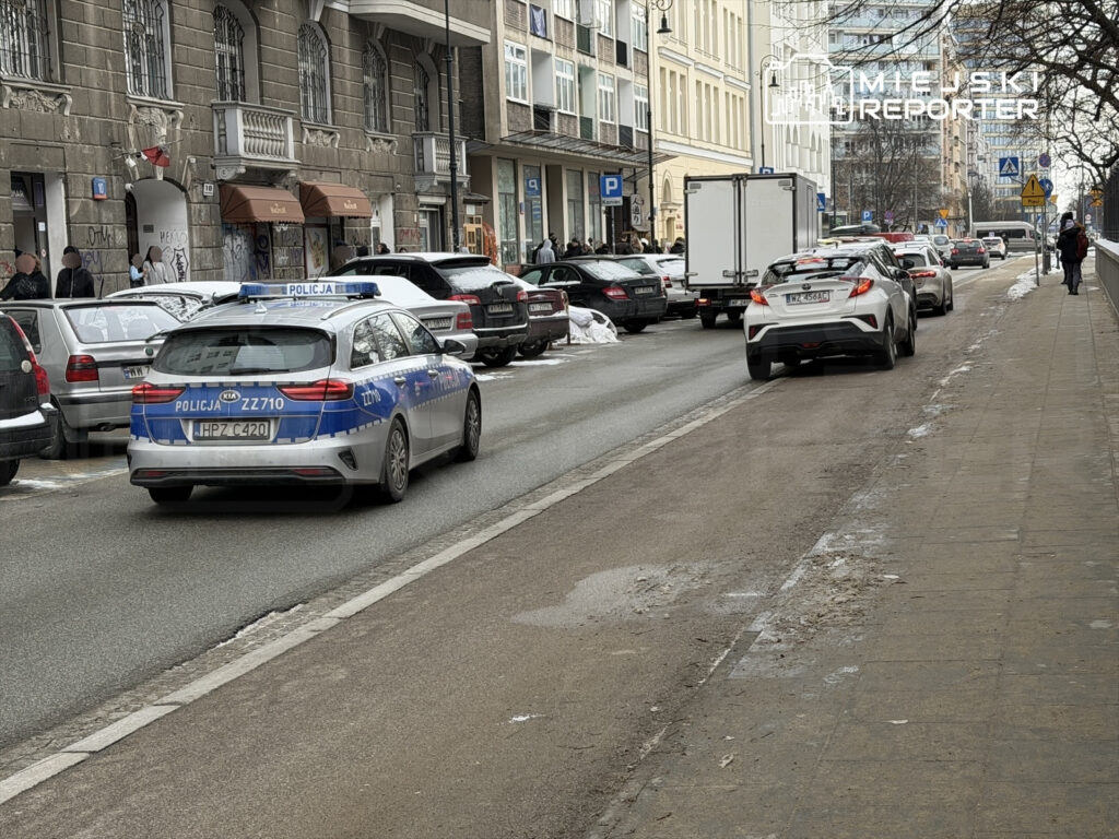 Policyjny radiowóz przejeżdża obok zaparkowanych samochodów na miejskiej ulicy w zimowej scenerii.