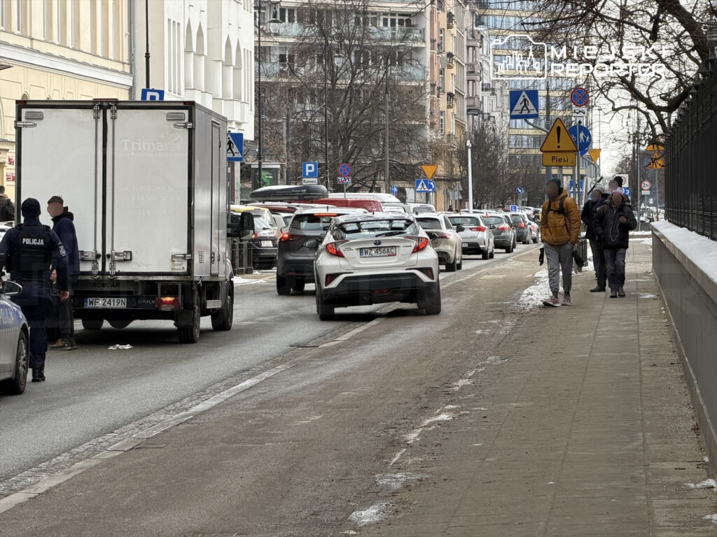 Policjant rozmawia z kierowcą dostawczego auta na zatłoczonej ulicy, obok przechodnie idą chodnikiem.