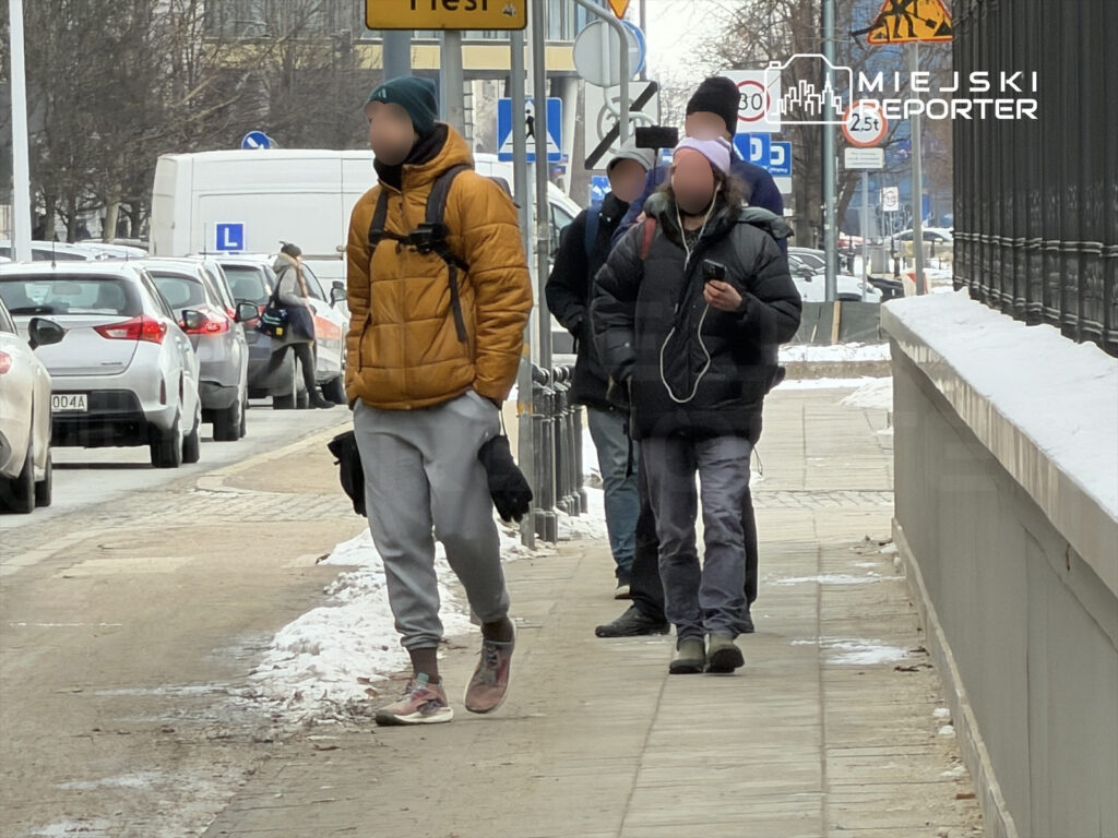 Grupa trzech mężczyzn idzie chodnikiem w mieście, mijając zaparkowane samochody i znaki drogowe.