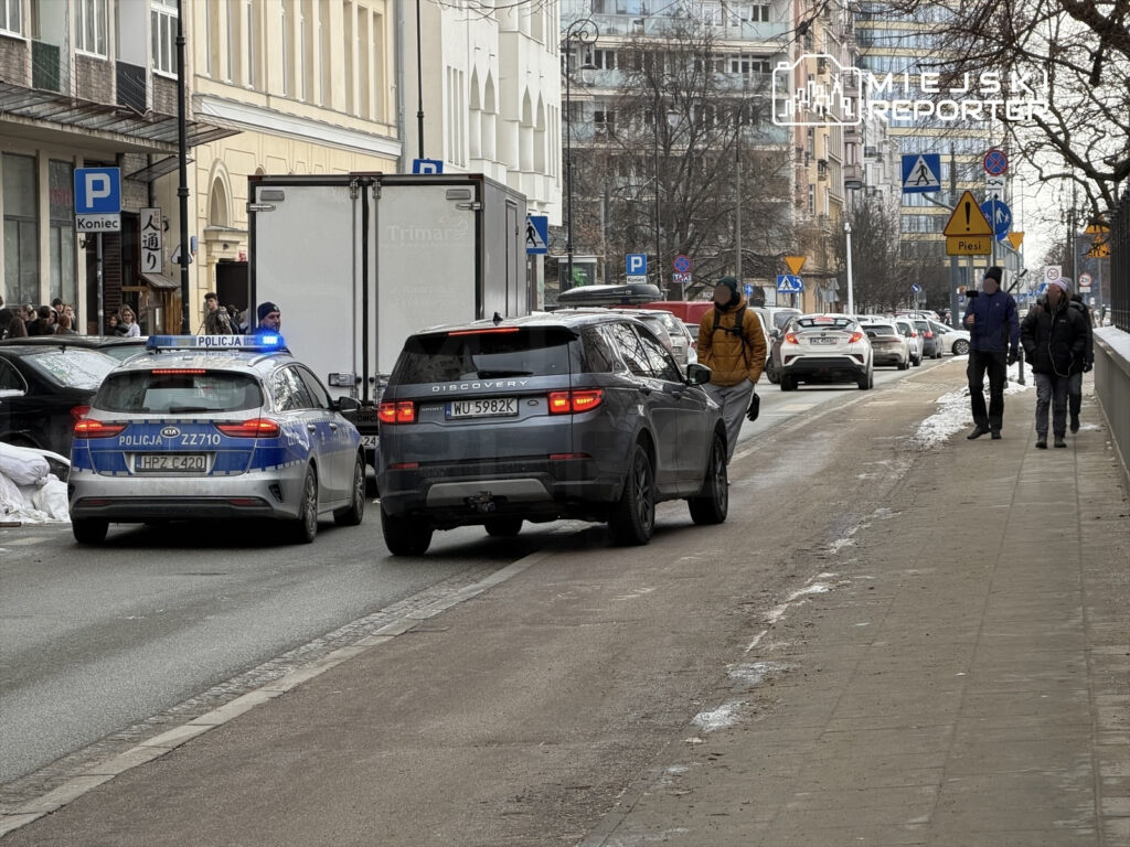 Policyjny radiowóz z włączonymi sygnałami świetlnymi zatrzymuje się obok dostawczego samochodu na ruchliwej ulicy.