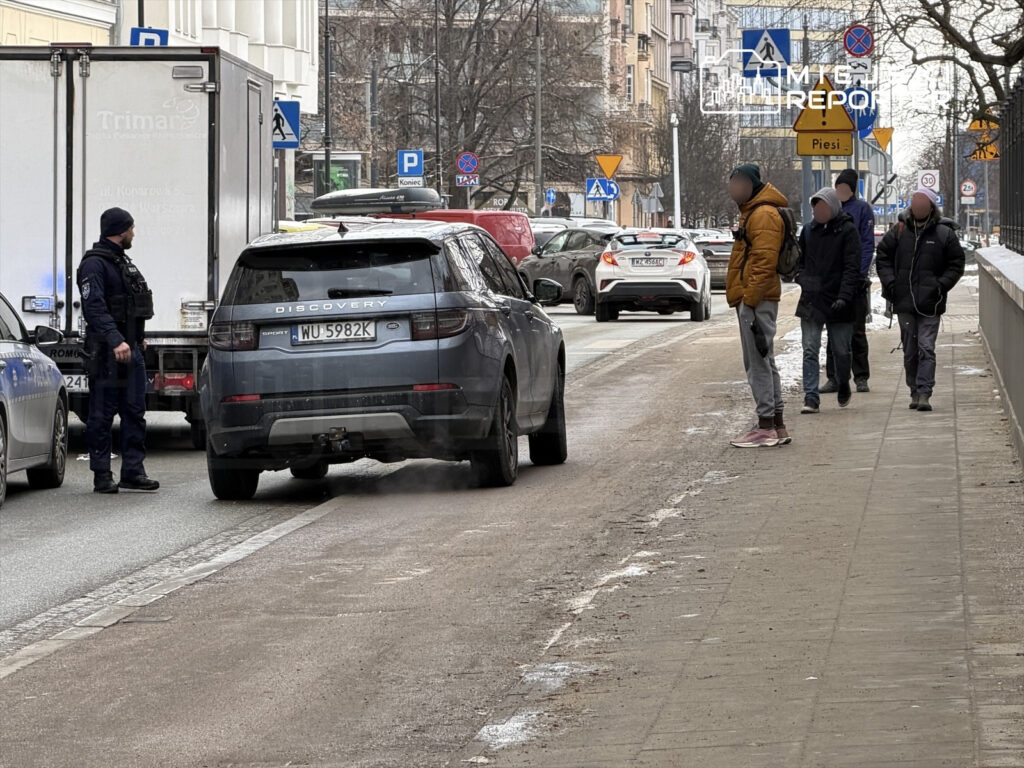 Policjant rozmawia z kierowcą SUV-a na ulicy, obok przechodzą piesi w zimowych kurtkach.
