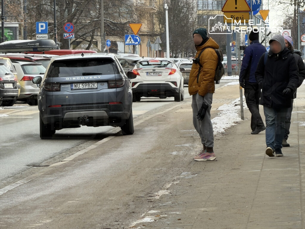 Mężczyzna w pomarańczowej kurtce stoi na chodniku obok zaparkowanego samochodu SUV, a w tle przechodzą inni piesi.