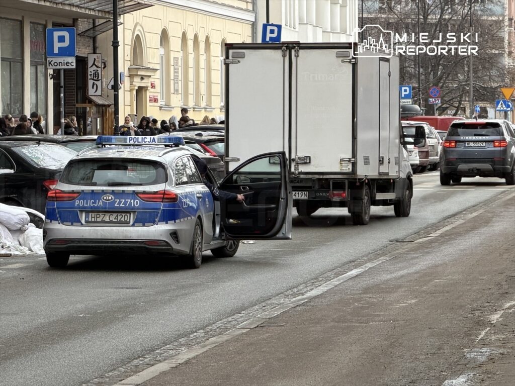 Policyjny samochód zaparkowany na ulicy z otwartymi drzwiami, obok zatrzymany dostawczy pojazd w miejskim otoczeniu.