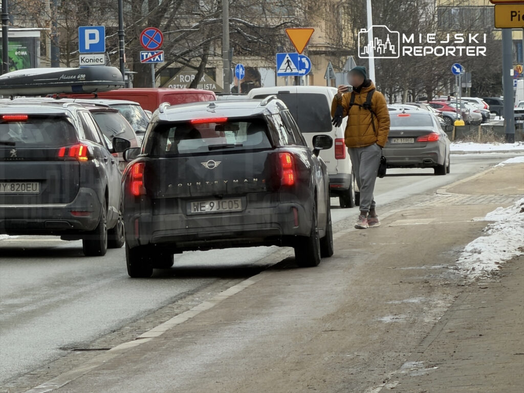 Kierowca w czarnym samochodzie zatrzymany na ruchliwej ulicy, obok przechodzi mężczyzna w żółtej kurtce, korzystający z telefonu.