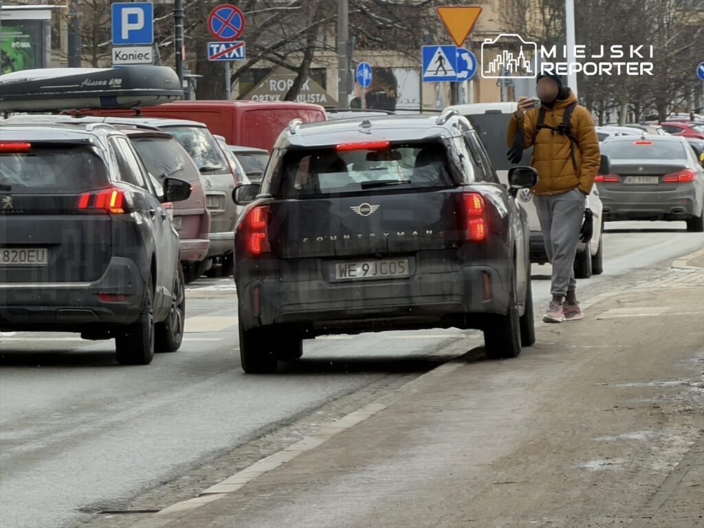 Mężczyzna w żółtej kurtce stoi na chodniku, trzymając telefon, obok przejeżdżające samochody w miejskim otoczeniu.