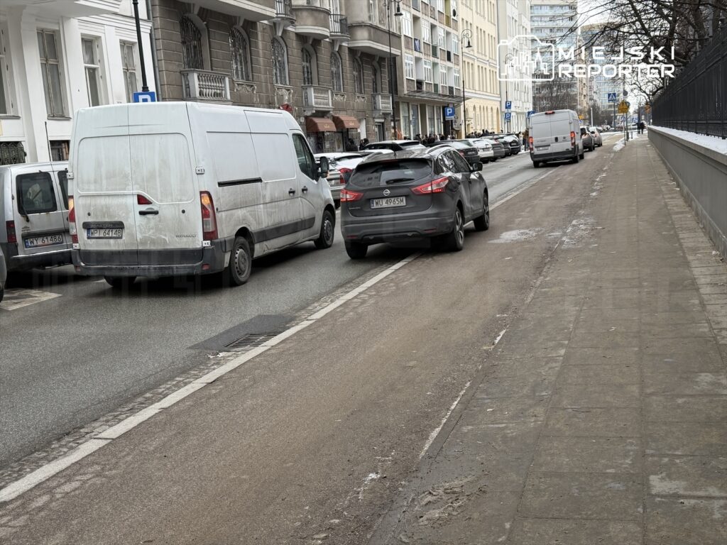 Na zatłoczonej ulicy dwa samochody dostawcze stoją w korku, a obok znajduje się chodnik z resztkami śniegu.