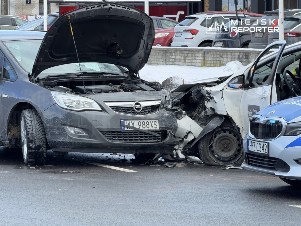 Dwa uszkodzone samochody osobowe na parkingu, jeden z otwartą maską, obok policyjny radiowóz.