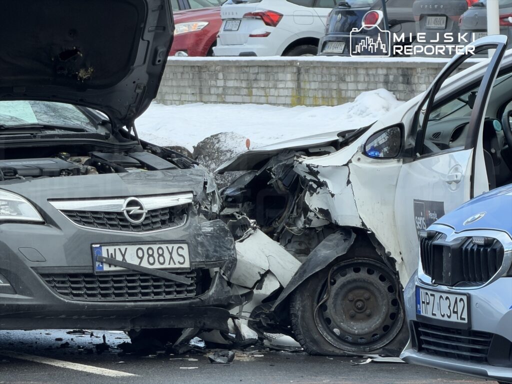 Zderzenie dwóch samochodów osobowych na parkingu, z widocznymi uszkodzeniami przodu jednego z pojazdów.