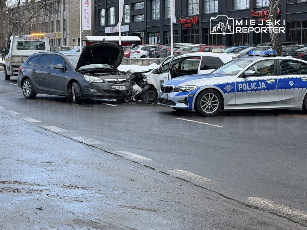 Kolidujące pojazdy osobowe na drodze, obok zaparkowany radiowóz policji oraz laweta.