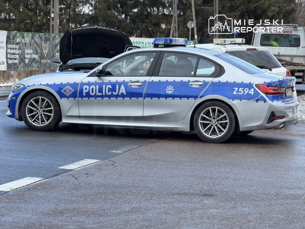 Policyjny radiowóz z niebieskimi oznaczeniami stoi na drodze, obok otwartej maski innego pojazdu.