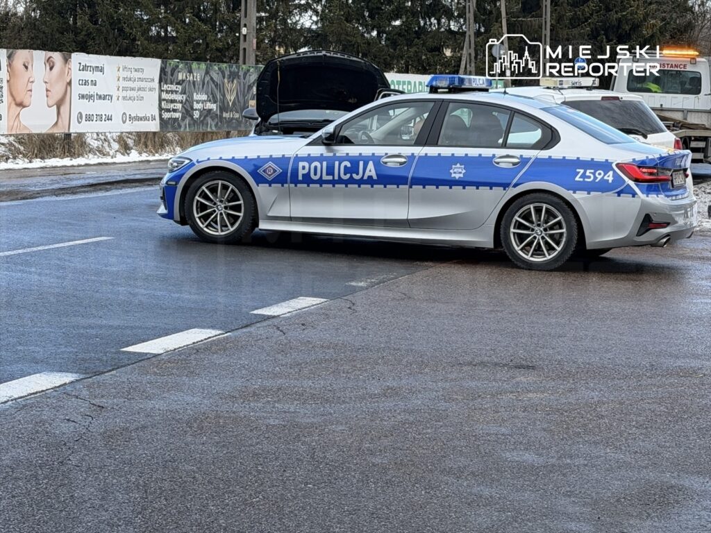 Policyjny radiowóz z otwartą maską stoi na drodze, obok widoczne reklamy na płocie.