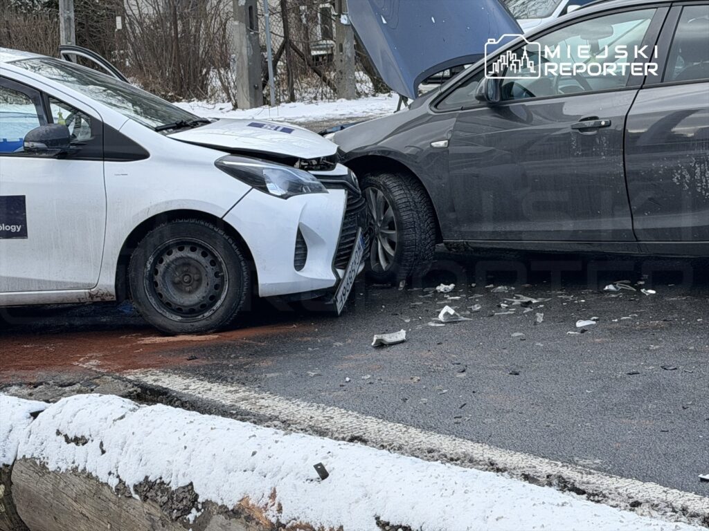 Zderzenie dwóch samochodów osobowych na drodze, jeden z pojazdów ma uszkodzoną maskę, wokół rozbite elementy.