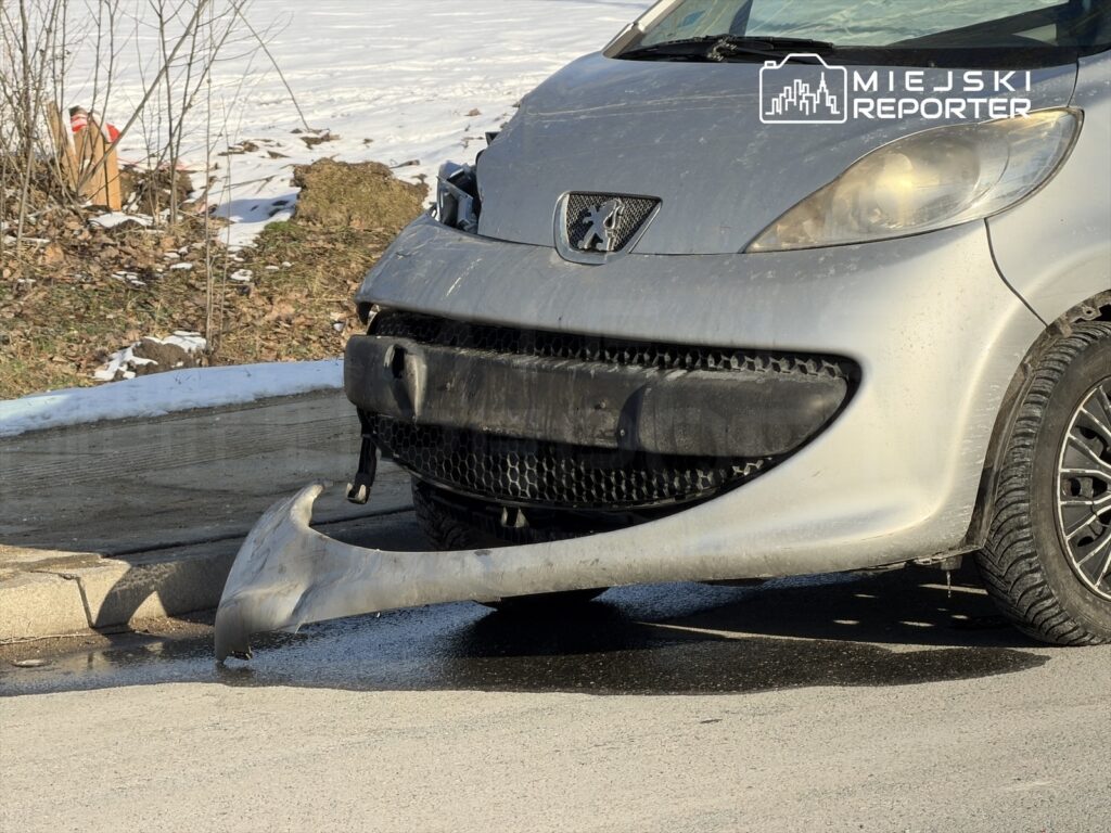 Uszkodzony samochód osobowy z odczepionym zderzakiem stoi na drodze, obok leżą resztki śniegu i liści.