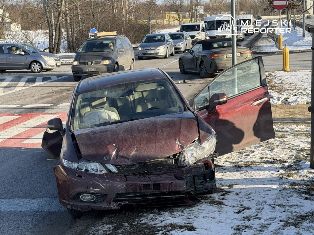 Czerwony samochód osobowy z uszkodzoną przednią częścią stoi na poboczu drogi, obok otwarte drzwi. W tle inne pojazdy w ruchu.