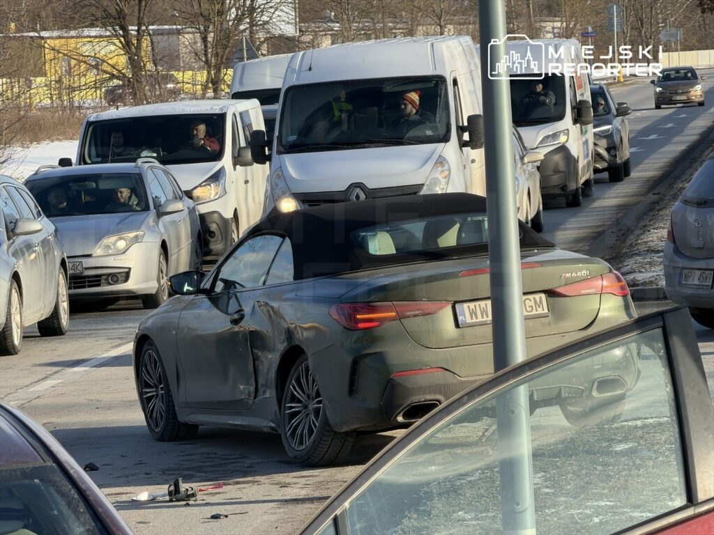 Zniszczony samochód sportowy zaparkowany na poboczu drogi, obok stojące pojazdy w korku.