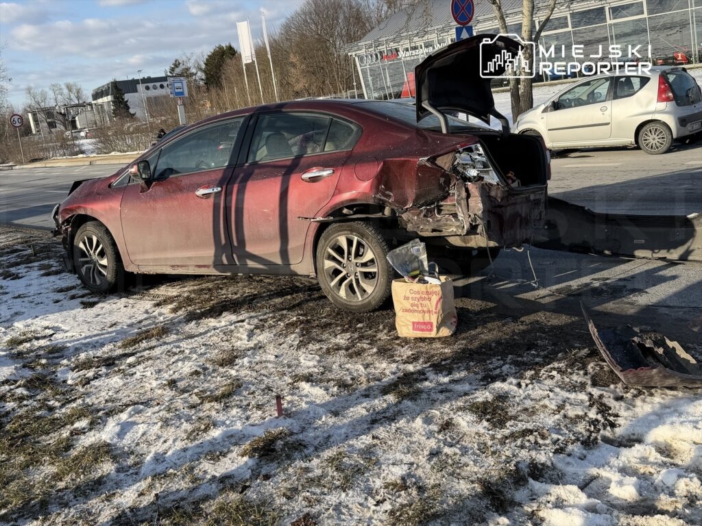 Uszkodzony czerwony samochód zaparkowany na poboczu, z otwartym bagażnikiem, obok leży torba z tekstem "Coś czuję ze siebie ze względu na..." w zimowym otoczeniu.