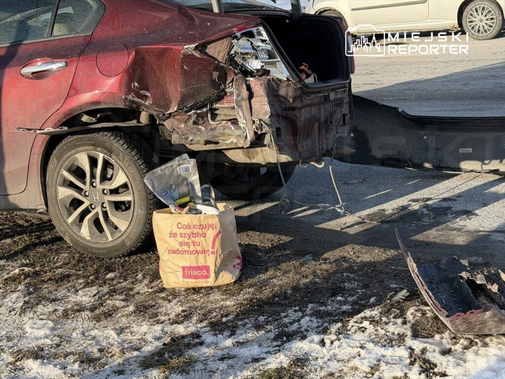 Czerwony samochód osobowy z uszkodzoną tylną częścią stoi na ośnieżonym parkingu, obok leży torba z napisem „Coś czuję, że szybko się tu zadomowię”.