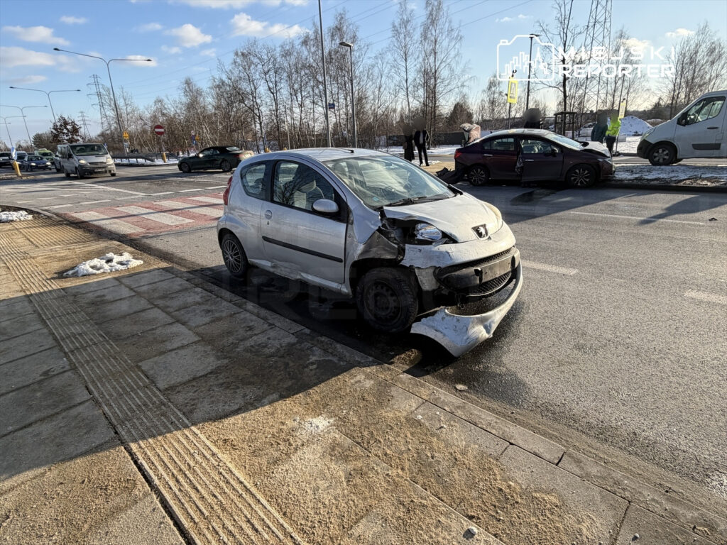 Uszkodzony samochód osobowy zaparkowany na poboczu drogi, obok ruchliwej skrzyżowania, z widocznymi śladami kolizji.