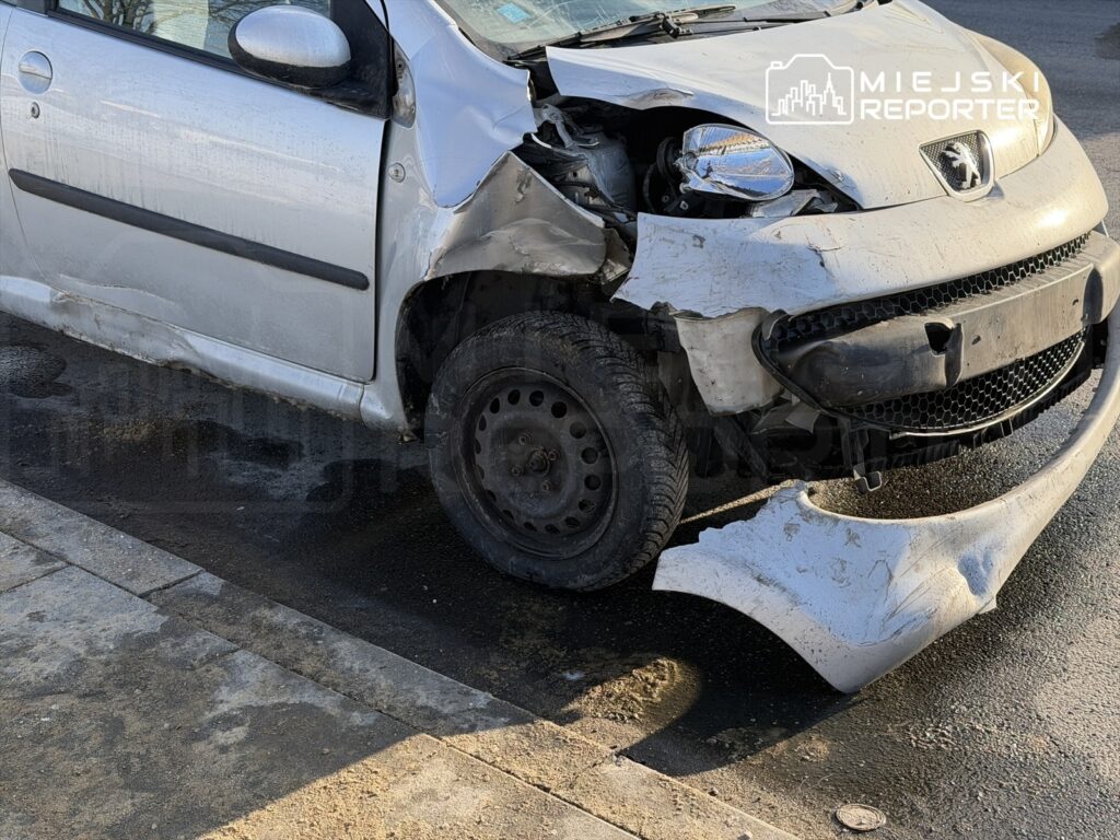 Uszkodzony samochód osobowy na poboczu drogi, z wgniecionym przodem i odłączonym zderzakiem.